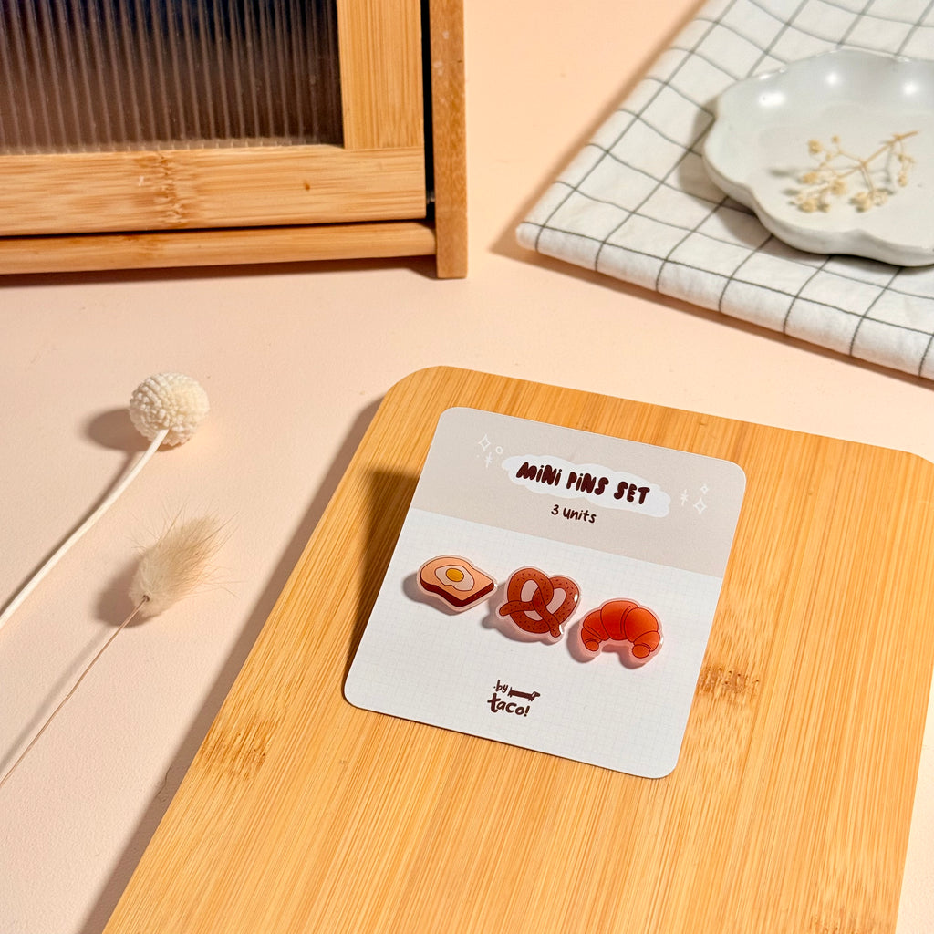 A mini pin set displayed on a white backing card, placed on a light wood tray. The set includes three small pins: a fried-egg shape, a pretzel and a croissant, all in warm orange and cream tones.