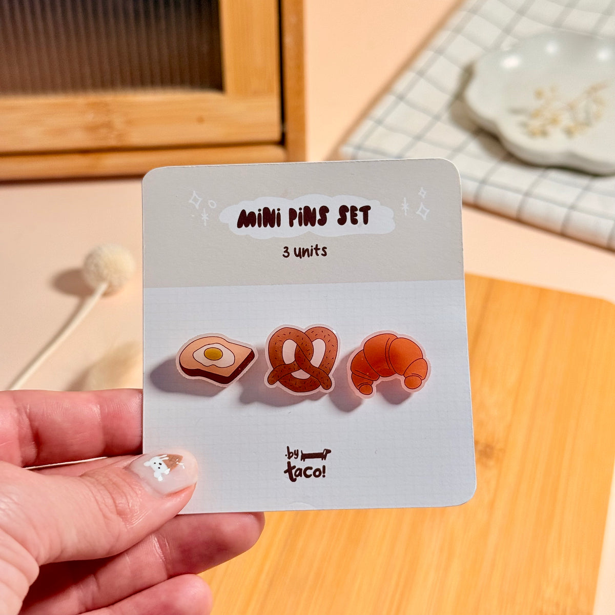 A mini pin set displayed on a white backing card, placed on a light wood tray. The set includes three small pins: a fried-egg shape, a pretzel and a croissant, all in warm orange and cream tones.