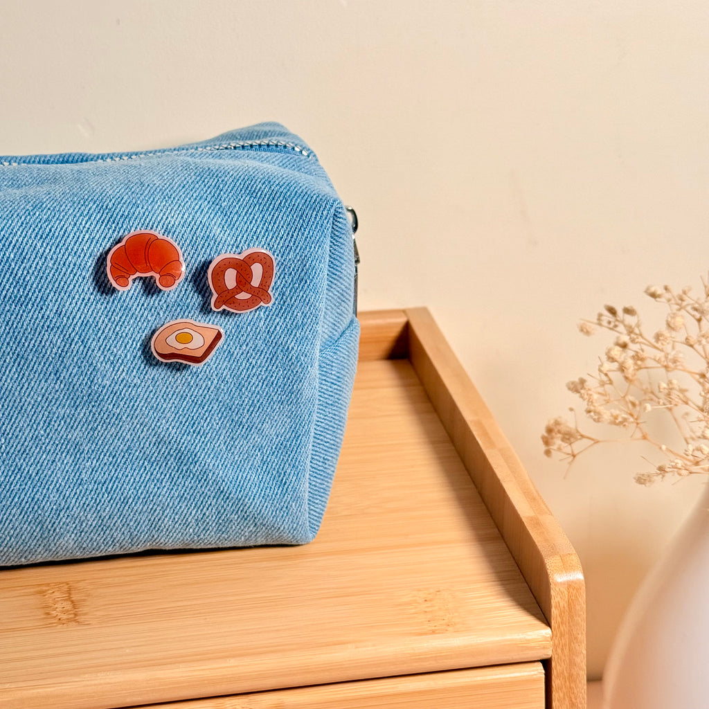 A light blue denim zippered pouch with three small food-themed enamel pins (croissant, pretzel, and egg) resting on a light wood surface against a warm beige background.